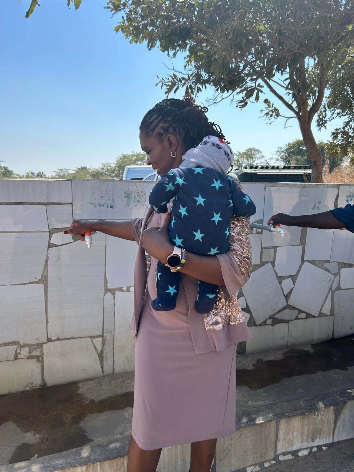 A mother holding her baby while drawing clean water from a tap at a community well